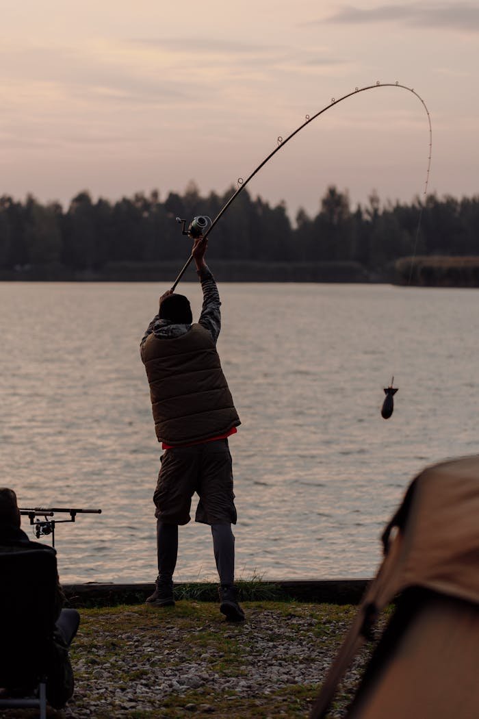 Silhouetted fisherman casting a rod by a tranquil lake at sunset.